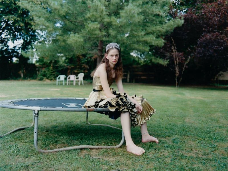 Barefoot woman in golden dress sits on trampoline in backyard. Lush green grass, trees, and white plastic chairs in background.