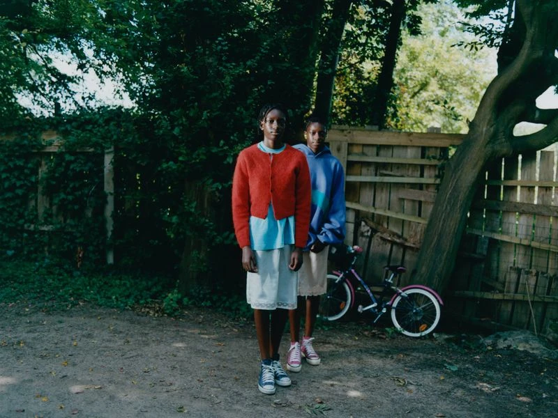 Two young girls stand in a yard with a bicycle in the background. The girl on the left wears a red cardigan. The girl on the right wears a blue sweatshirt.