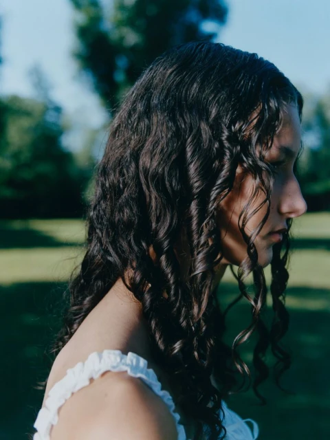 Profile of a young woman with long, curly dark hair wearing a white, frilled top in a natural outdoor setting with blurred green background.