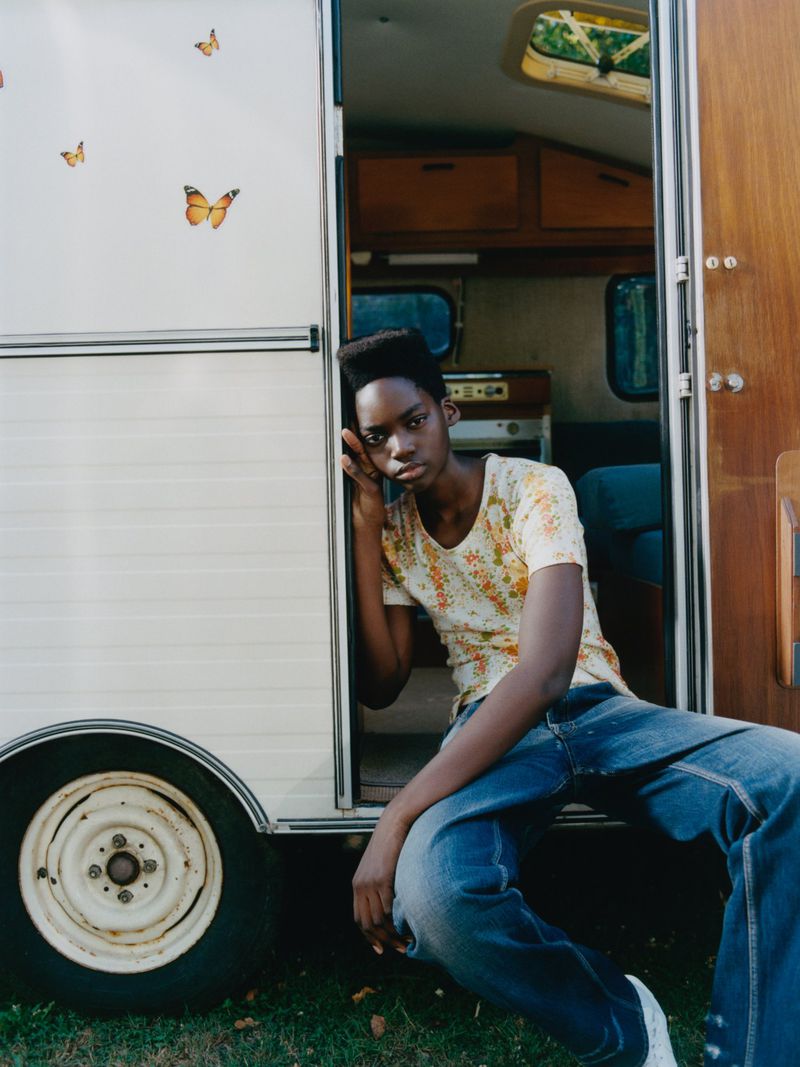 Young woman with dark hair and denim jeans sits in the doorway of a vintage camper with butterfly decals on the exterior.