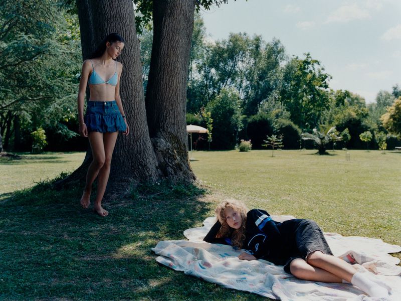 Two women in casual summer clothing are in a sunny park with large trees, grassy lawn, and foliage; one woman lies on a blanket.