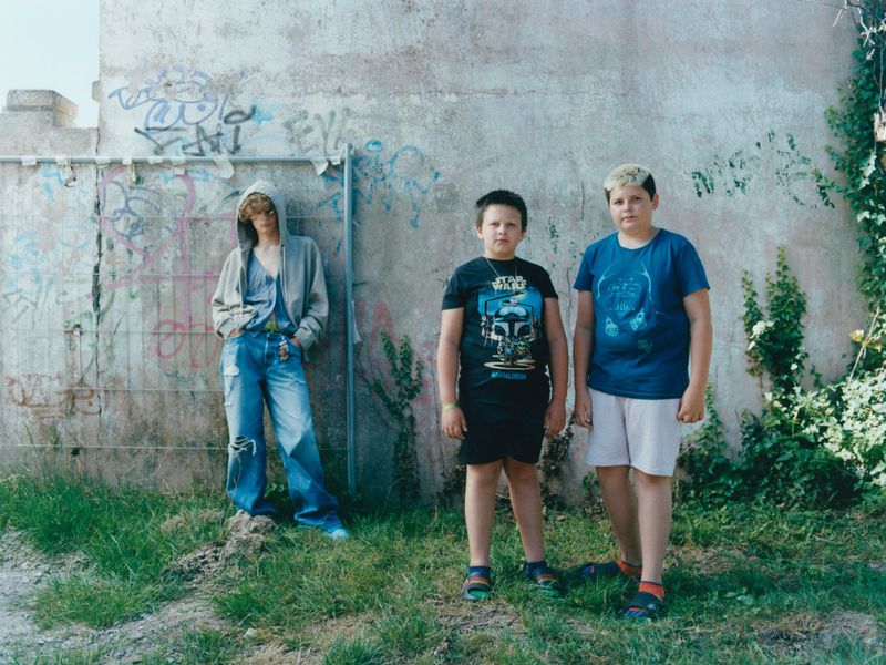 Three children stand in front of a graffiti-covered wall next to greenery; one child wears a hoodie and the other two wear graphic tees.