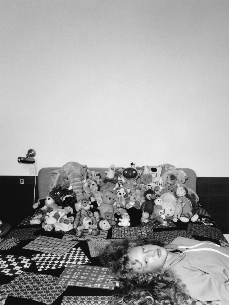Person lying on a bed covered in patchwork quilt with stuffed animals piled up on the headboard against a white wall in black and white.