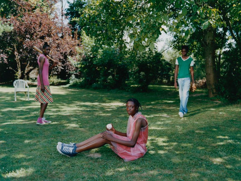 Three Black women in a grassy yard, one sitting with a baseball, another holding a bat, and the third with a baseball glove.