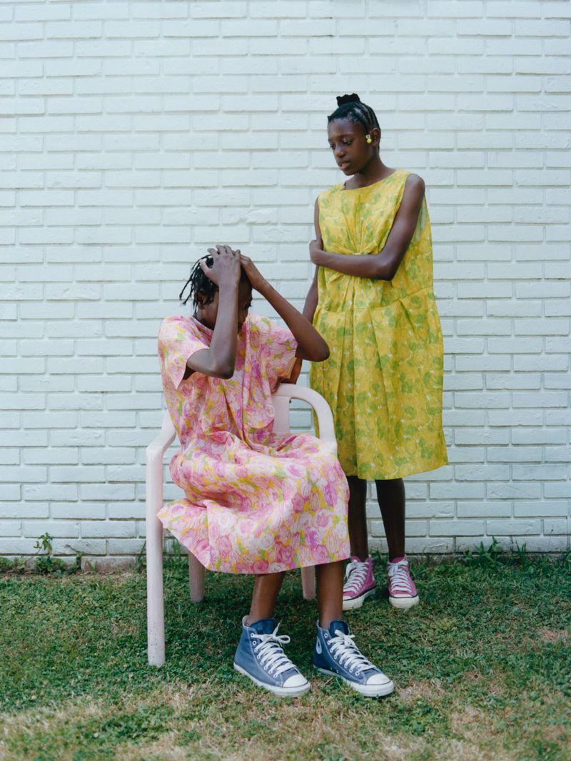 Two African-descent women outside with a white brick wall background, one sitting on a chair and one standing in yellow dress.