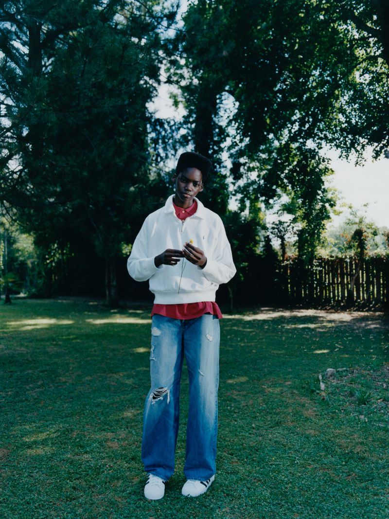 Full shot of a young adult Black woman standing on grass in ripped jeans, a red shirt, and white jacket under green trees.