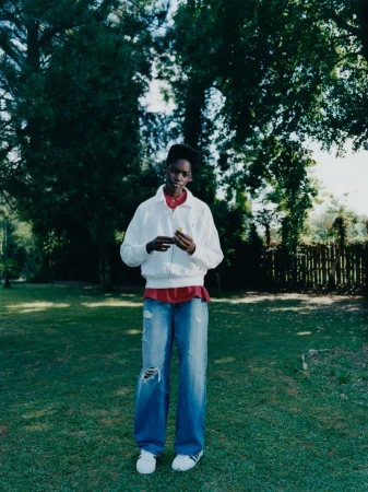 Full shot of a young adult Black woman standing on grass in ripped jeans, a red shirt, and white jacket under green trees.