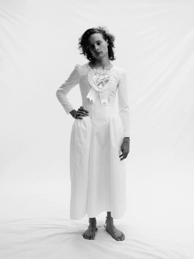 Person in white dress with heart-shaped decoration poses barefoot in a photo studio with bright, even lighting and neutral background.
