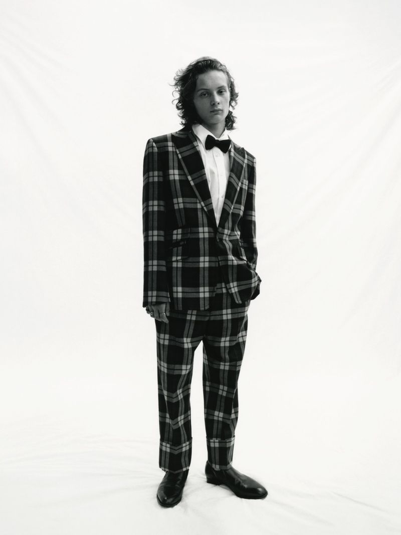 Young adult stands in a plaid suit with a bow tie, captured in a full shot against a bright backdrop. The attire suggests formal fashion.