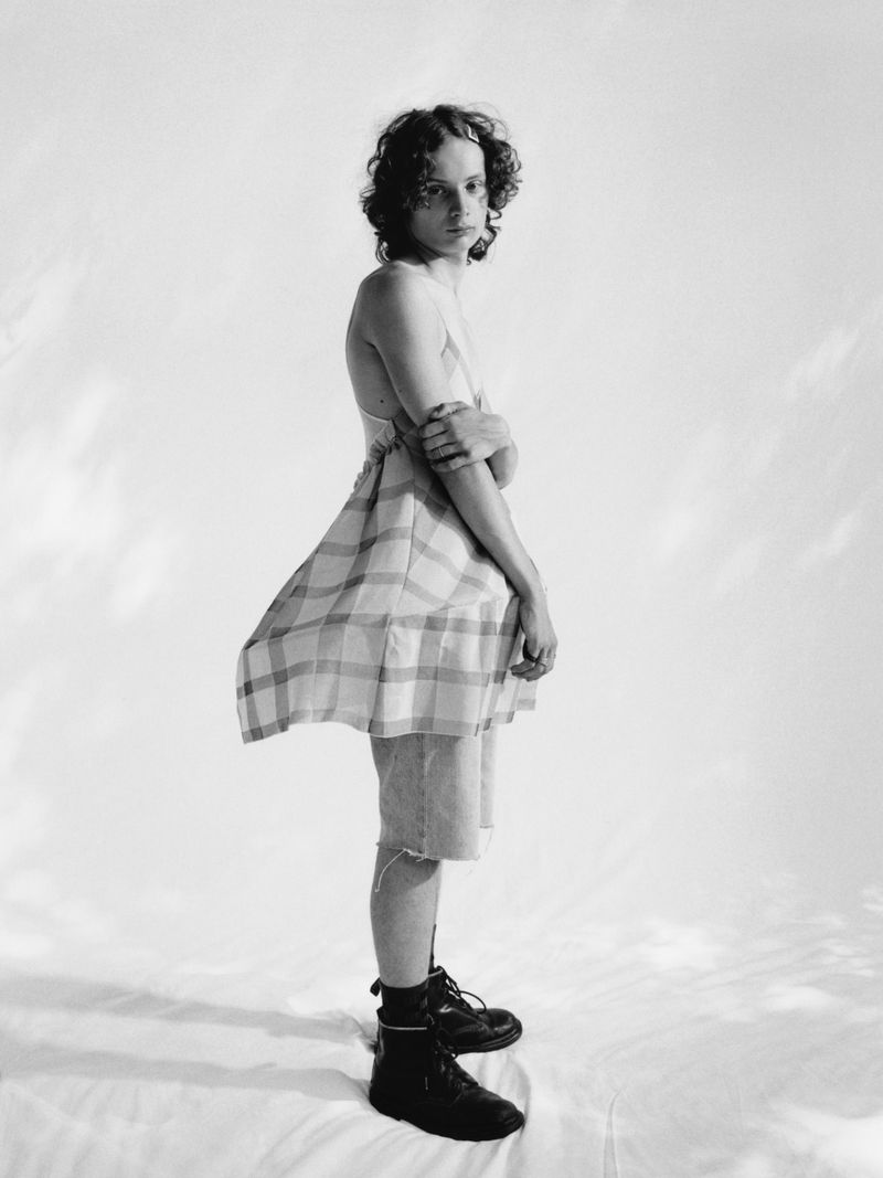 Monochrome portrait of an androgynous person in plaid fabric, jean shorts, and boots against a white backdrop in a photo studio.