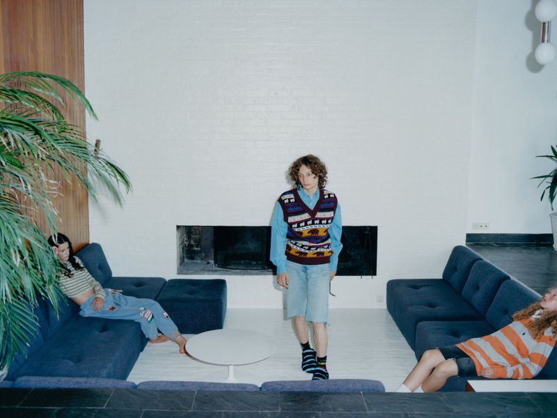 Three people relax in a modern living room with blue couches, white walls, and a large houseplant. Casual fashion and interior design.