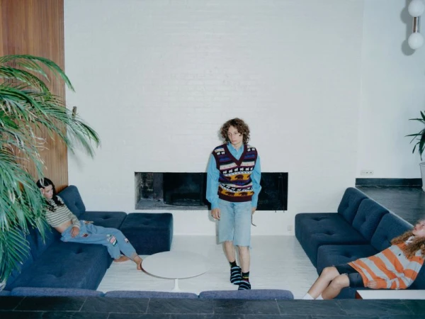 Three people relax in a modern living room with blue couches, white walls, and a large houseplant. Casual fashion and interior design.