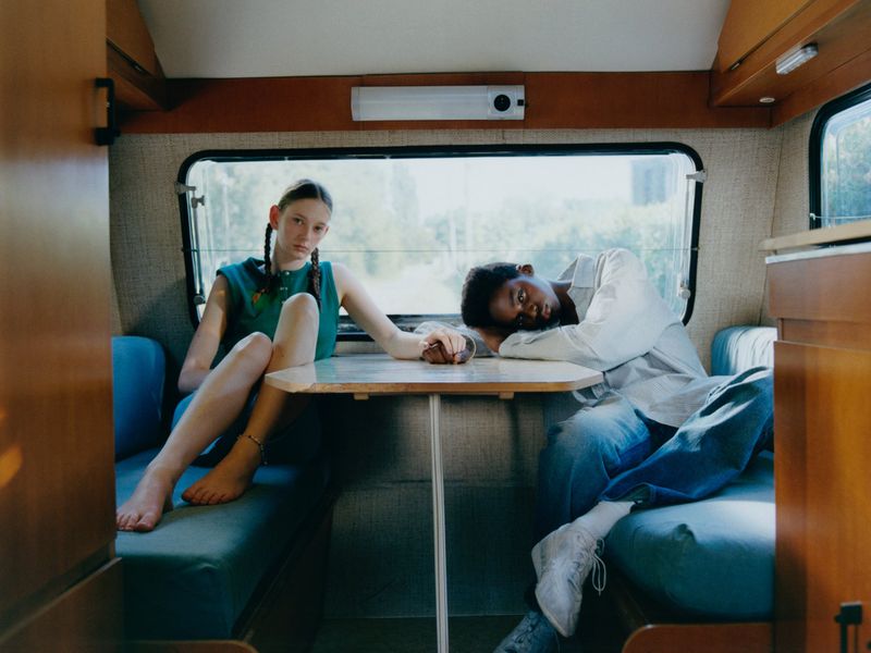 Two young women sit at a small table in a camper van. One woman rests her head on the table, and they are holding hands.
