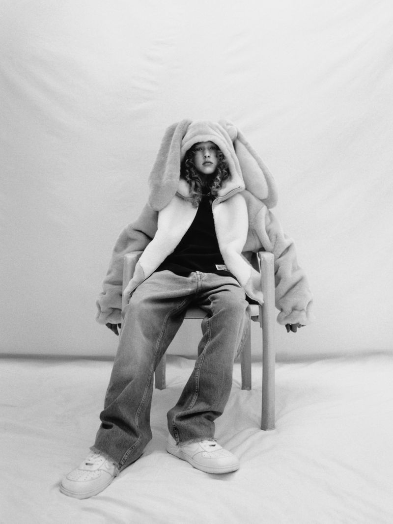 Child sits in studio wearing faux-fur rabbit coat and light-wash jeans, illuminated by soft lighting against a neutral backdrop.