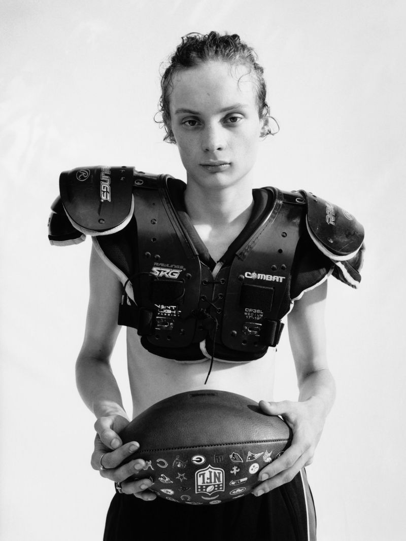 Studio portrait of a young adult holding a football, wearing American football shoulder pads, in black and white, against a white backdrop.