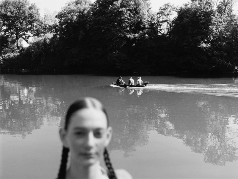 Monochrome photograph of a woman in the foreground, out of focus, and a boat with a group of people on a river surrounded by trees.