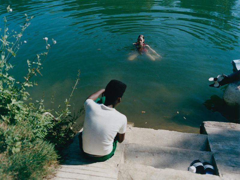 Woman swims in a green lake, viewed from concrete steps where a man sits and another rests, with abundant green foliage visible.