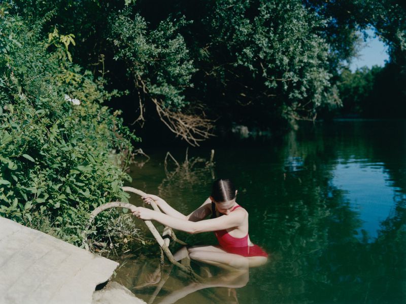 A woman in a red swimsuit sits in a river surrounded by green trees. The woman is holding a curved tree branch with both hands.
