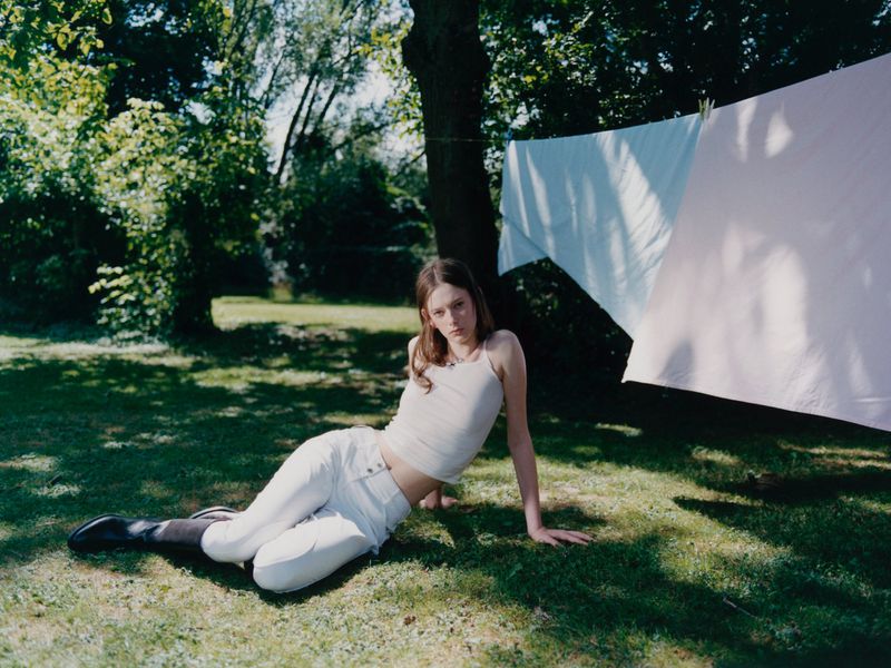 A young woman in white jeans and top poses on grass in a bright outdoor setting, with white sheets hanging on a clothesline in the background.