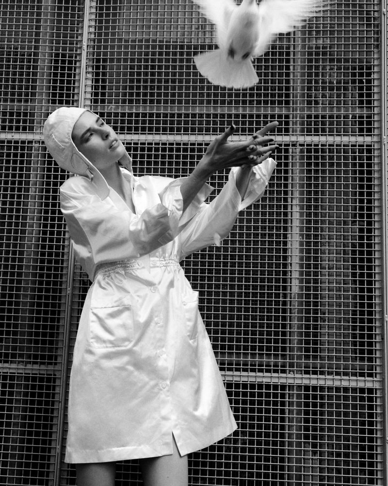 Monochrome image of a woman in a white robe releasing a white dove, set against a geometric metal grid backdrop in an indoor setting.