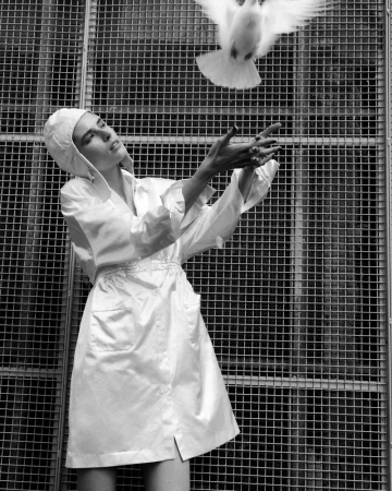Monochrome image of a woman in a white robe releasing a white dove, set against a geometric metal grid backdrop in an indoor setting.