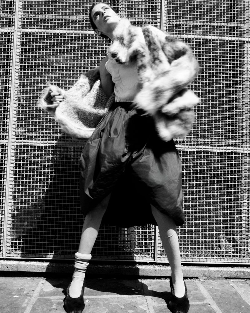 Monochrome fashion shot of a woman in a fur coat and skirt, posing in front of a metal grate, with dramatic lighting and unique style.