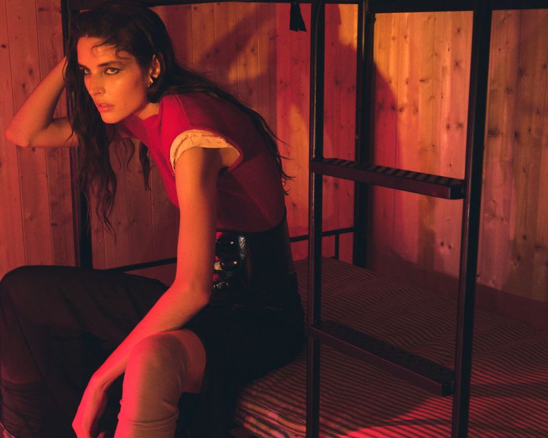 A stylish woman with dark hair is sitting on a metal bunk bed frame in front of a rustic wood wall bathed in red and orange light.