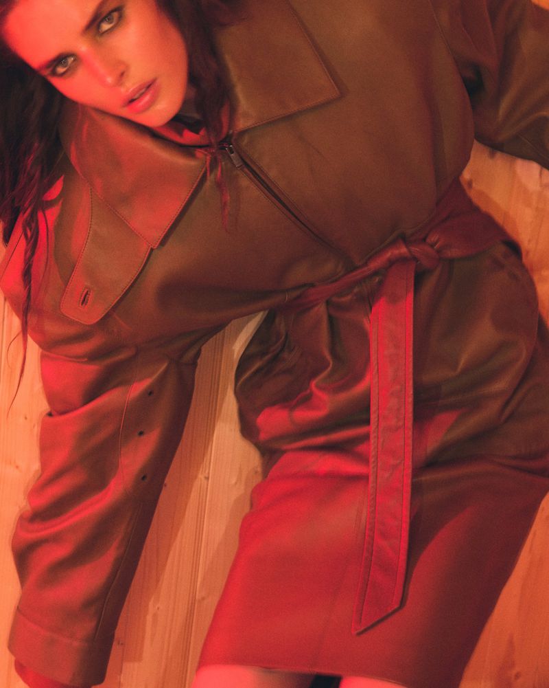 Brunette woman in a brown belted coat and matching skirt is lying on a wooden floor, illuminated by a red light.
