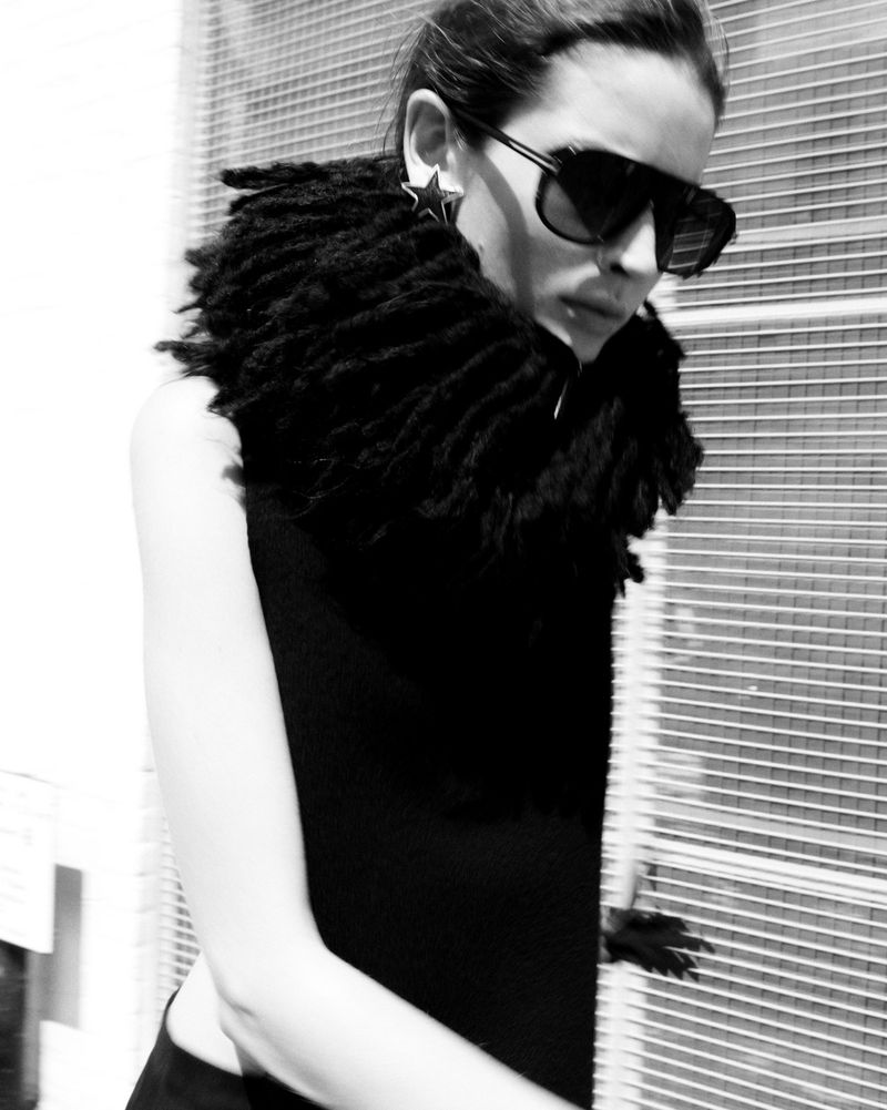A woman wearing sunglasses and a large fringed collar poses against a metal fence in a black and white fashion shot.