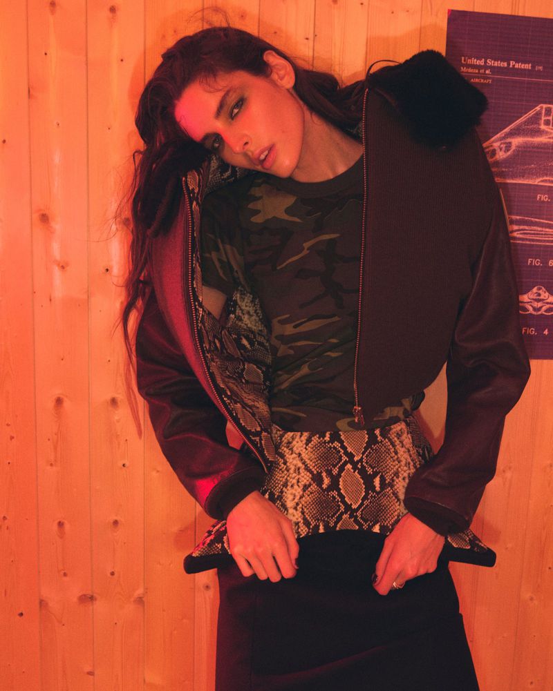 A brunette woman with long dark hair poses indoors wearing a camo t-shirt, snakeskin skirt, and dark jacket in an orange-lit environment.