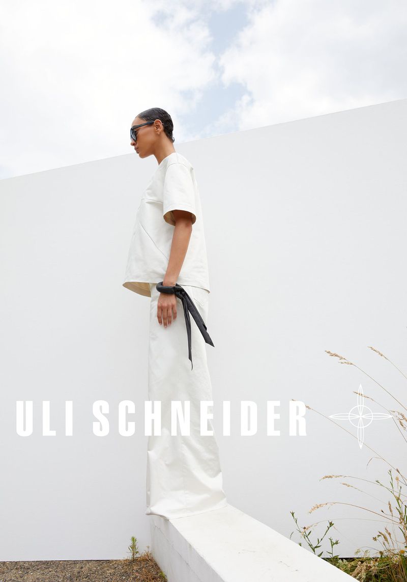 A woman in a white outfit with sunglasses and wrist cuff stands outside against a white wall under a bright sky, showcasing a fashion editorial.