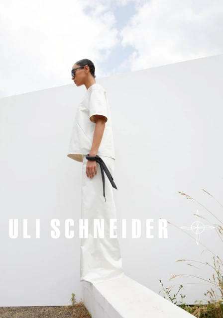 A woman in a white outfit with sunglasses and wrist cuff stands outside against a white wall under a bright sky, showcasing a fashion editorial.