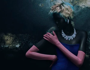 Model wearing long pink opera gloves, a blue strapless dress, and an ornate crystal necklace against a dark textured background.