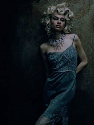 Woman in a green lace slip dress with blonde curly hair and an ornate necklace posing against a dark textured wall