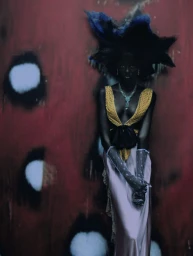 Black model wearing a dramatic dark sculptural headpiece, yellow plunging top, and lavender satin skirt against a red textured backdrop.