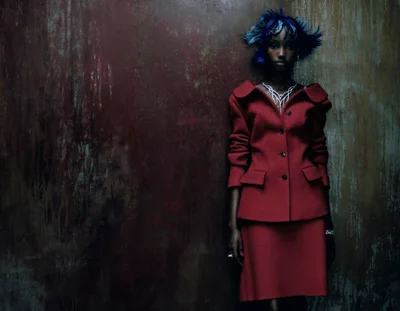 Model with sculptural blue hair wearing a red skirt suit and ornate necklace against a dark textured wall.