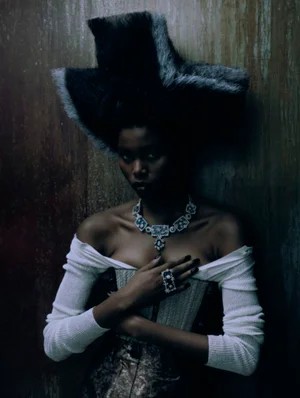 Model wearing a sculptural dark fur headpiece, off-the-shoulder white top, corset, and ornate statement necklace against a wooden wall.