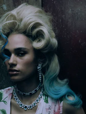 Close-up portrait of a model with voluminous blonde and blue-tipped hair wearing sparkling diamond jewelry and floral top.