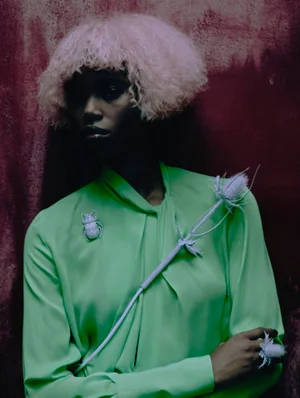 Black model with textured pink hair wearing a vivid green blouse and a beetle brooch against a dark textured burgundy backdrop.