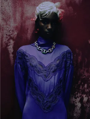 Model with sculpted white hair wearing a high-neck purple lace dress and chunky chain necklace against a textured crimson backdrop.