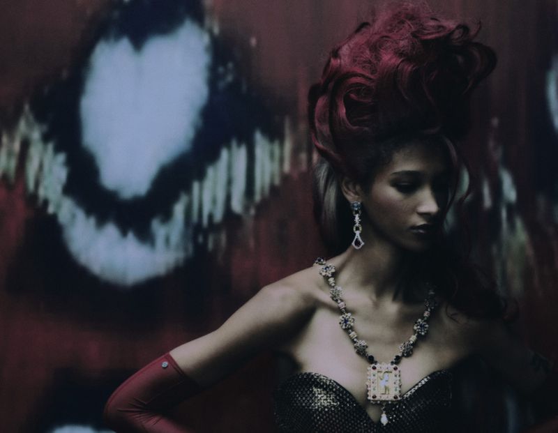 Adult female model poses wearing a strapless black dress, jewelry, and red gloves against a blurred red and black background in a studio.