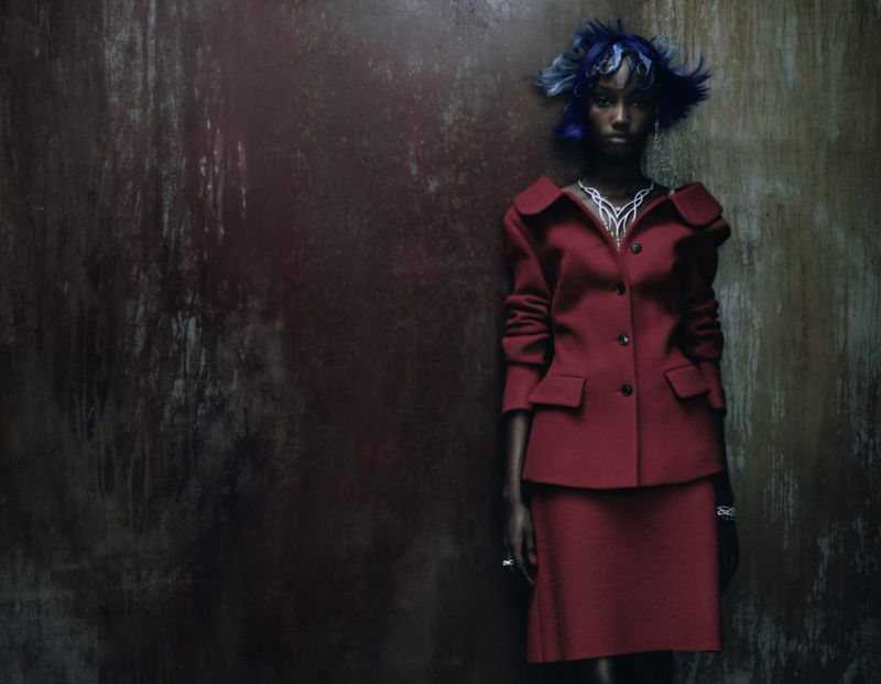 Adult female poses in red skirt suit against textured wall with blue hair. Fashion editorial shot with dramatic lighting in studio.