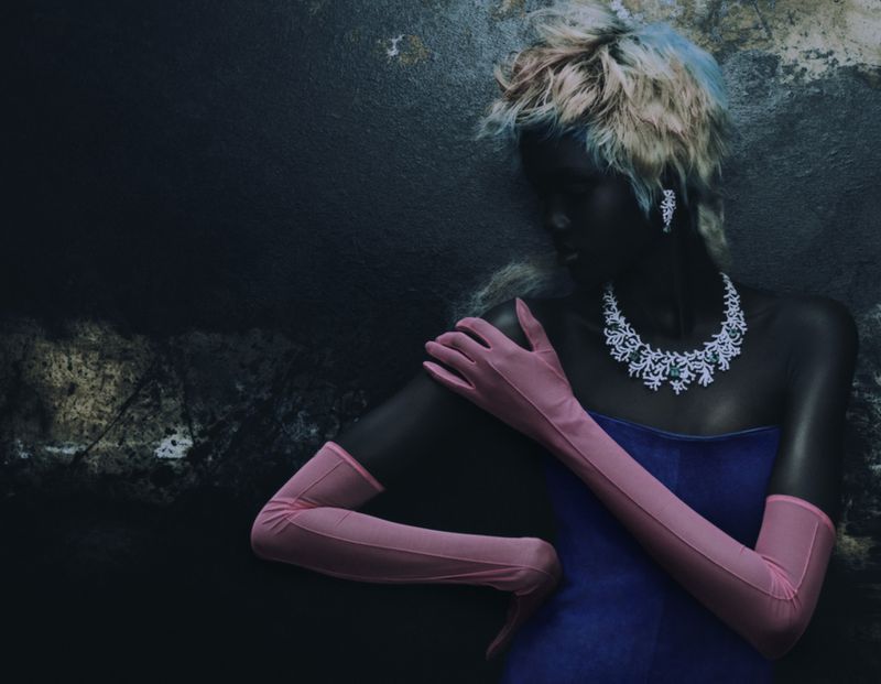 Elegant woman in a blue dress with jewelry and pink gloves poses against a dark textured wall in a fashion studio setting.