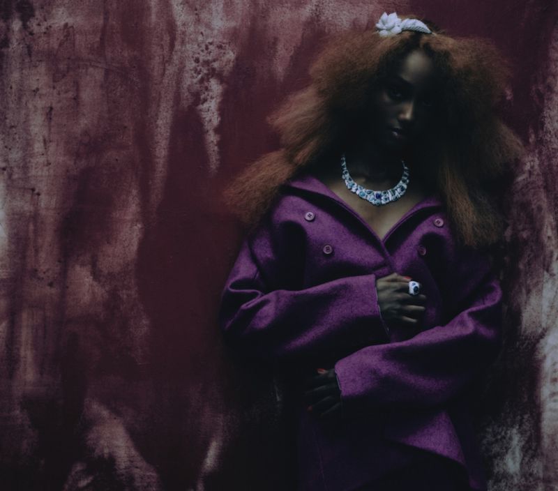 Woman in purple coat and statement necklace poses against a textured mauve backdrop in a beauty or fashion editorial photograph.