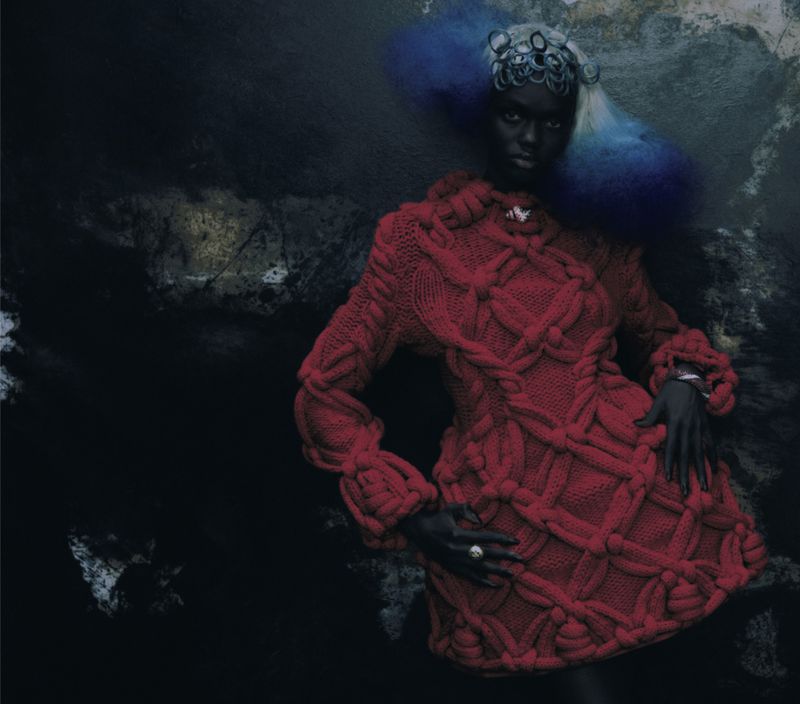 A model with dramatic blue and white hair poses in a red cable knit dress against a dark textured backdrop in a studio fashion shot.