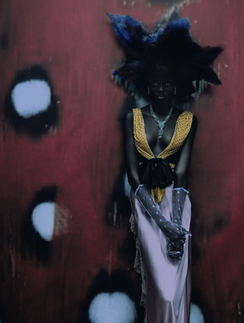 Woman in avant-garde fashion stands against a textured red backdrop with blurred white spots. She wears a couture dress and statement jewelry.
