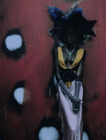 Woman in avant-garde fashion stands against a textured red backdrop with blurred white spots. She wears a couture dress and statement jewelry.