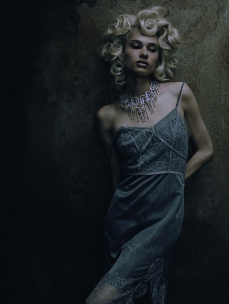 Blonde woman poses wearing a green dress and ornate jewelry, leaning against a mottled wall in a dimly lit indoor setting.