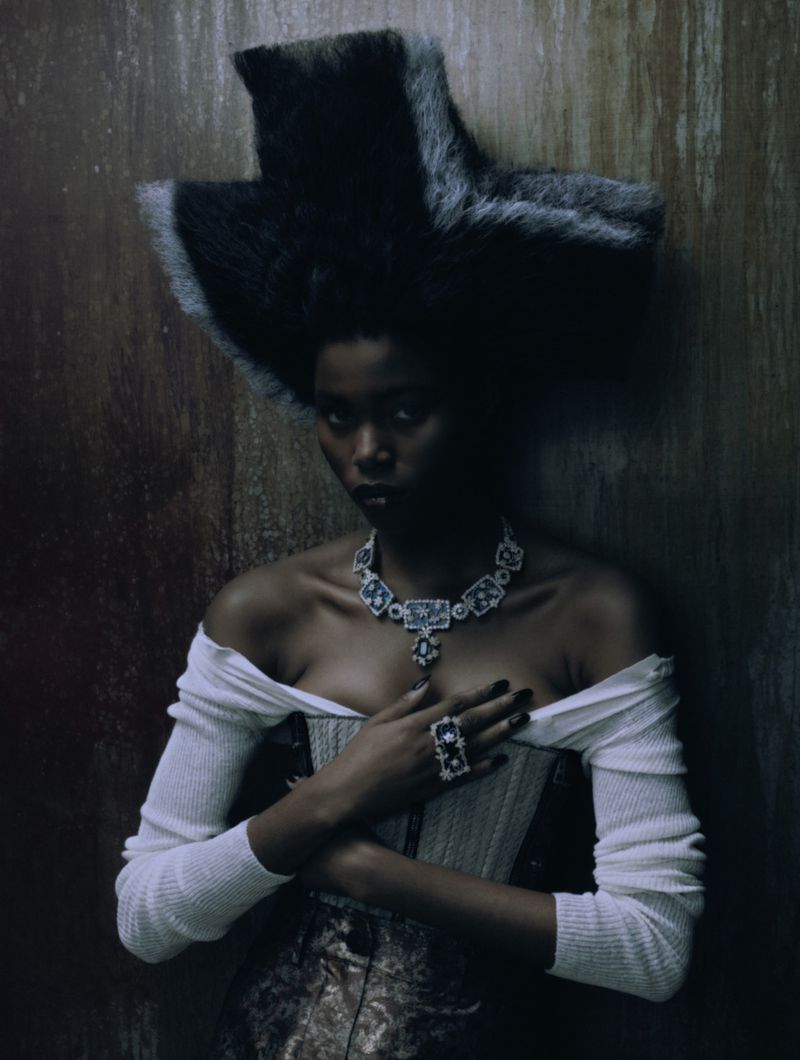 Elegant woman wearing large fur hat, diamond necklace, and corset poses in front of textured neutral wall in dark indoor setting.