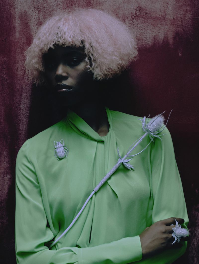 Dark-skinned woman poses against a red wall wearing a green blouse, insect brooch, white wig, and flower ring in a high-fashion setting.